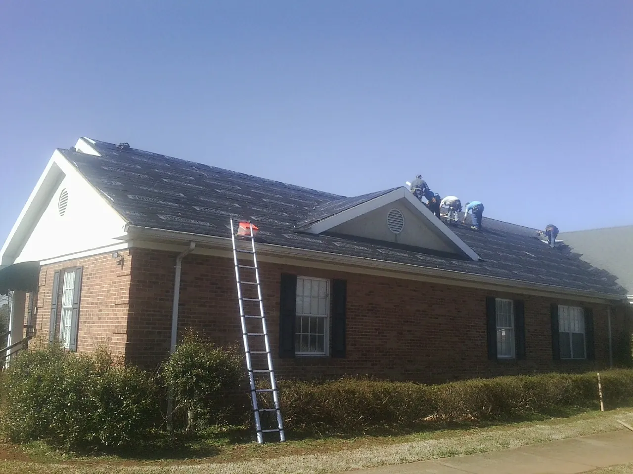 Crew tearing shingles off a brick ranch roof with a ladder set — Emergency Roof Repair work by our crew in Delong