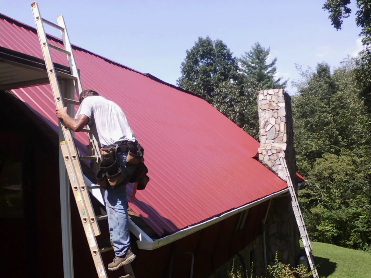 Roofer on a ladder beside a red metal roof and stone chimney — Soffit & Fascia Repair by Roofing Brothers in Delong